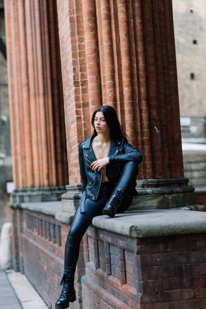 Портрет красивой молодой брюнетки женщина в стильной черной кожаной куртке, она улыбается на городском фоне, счастливое время, концепция путешествия. - Фото, изображение