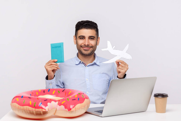 Cheerful man holding paper airplane and passport in hands, sitting in office workplace with rubber ring on desk, planning travel vacation, weekend trip. studio shot isolated on white background - Foto, imagen