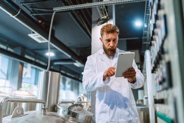  Professional caucasian handsome technologist expert in white uniform standing in pharmaceutical or food factory  - production plant checking productivity and quality using tablet computer. - Fotografie, Obrázek