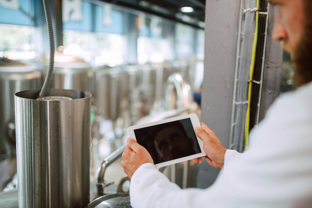  Großaufnahme eines Tablet-Computers in der Hand eines professionellen kaukasischen Technologen in weißer Uniform in der Produktionsanlage - Lebensmittel- oder Getränkefabrik - Foto, Bild