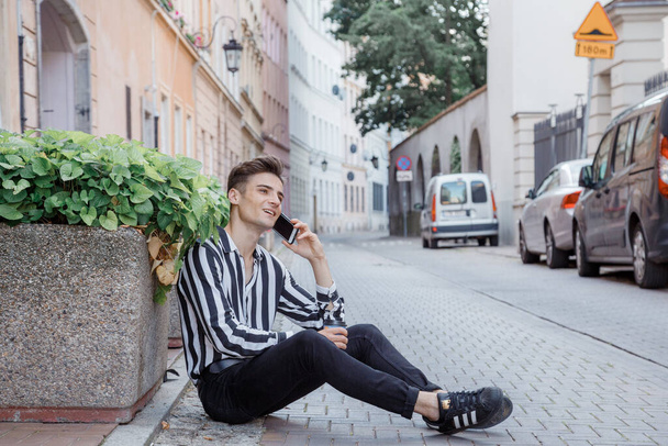 Ein junger Mann in gestreiftem Hemd sitzt auf der Straße in der Nähe eines Beetes mit rosa Blumen und telefoniert. Stilvoller junger Mann sitzt auf einer alten Straße in der Stadt. Unterwegs durch die Städte Europas. - Foto, Bild