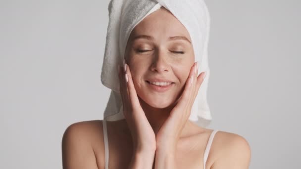 Młoda piękna kobieta z ręcznikiem na głowie szczęśliwie dotknąć jej oczyszczonej twarzy na szarym tle - Materiał filmowy, wideo