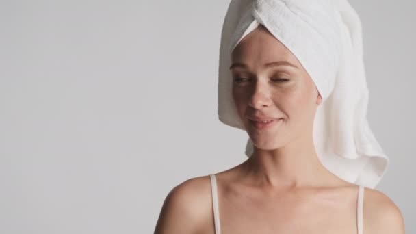 Młoda piękna kobieta z ręcznikiem na głowie patrząc na przestrzeń kopiowania i szczęśliwie dotknąć jej oczyszczoną twarz przed kamerą na szarym tle - Materiał filmowy, wideo
