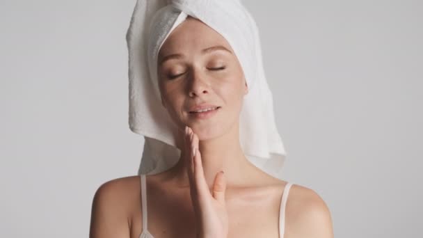 Mladá krásná žena s ručníkem na hlavě dotknout její očištěný obličej smyslně dívá do kamery přes šedé pozadí - Záběry, video