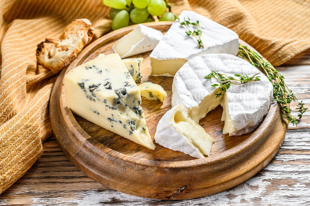 Assorted cheeses on a round wooden cutting Board. Camembert, brie and blue cheese with grapes. White wooden background. Top view. - Photo, Image