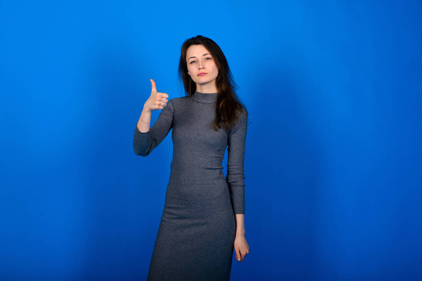 Photo of smiling, cheerful young female in grey dress on blue background. Emotional portrait - Photo, Image