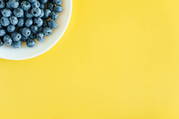 fruit and oat, vitamin, healthy food and drink, traditional medicine, gastronomy concept - layout of purple and blue blueberry berries in white plate or vessel on bright yellow background copy space - Photo, Image