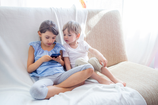 Χαριτωμένα μικρά αδέλφια έχουν μια καλή στιγμή στο σπίτι έννοια. Κοντά ο μικρός αδελφός και η αδελφή παίζουν με το tablet. Παιδιά κάθονται στον καναπέ, παίζοντας με το tablet. Τα παιδιά παίζουν ένα παιχνίδι σε ένα κινητό τηλέφωνο - Φωτογραφία, εικόνα