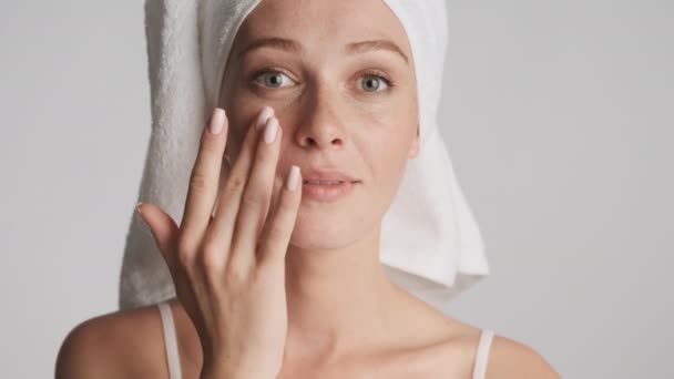 Retrato de mujer hermosa con toalla en la cabeza aplicando sensualmente crema hidratante en la cara sobre fondo gris
 - Imágenes, Vídeo