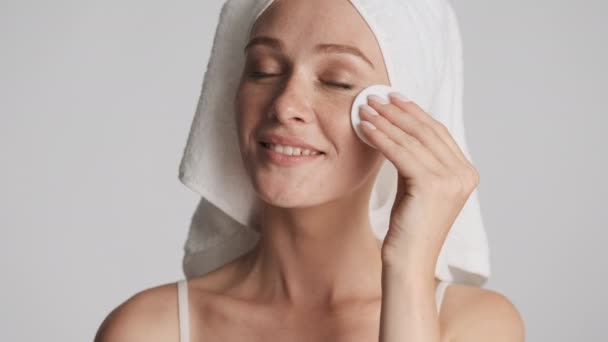 Όμορφο κορίτσι με πετσέτα στο κεφάλι καθαρισμένο πρόσωπο με σφουγγάρι βαμβάκι πάνω από το γκρι φόντο - Πλάνα, βίντεο