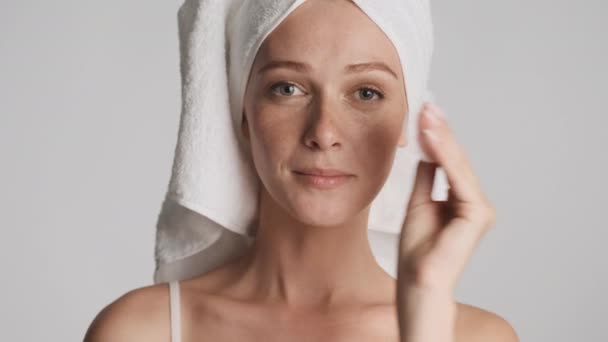 Νεαρή ελκυστική γυναίκα με πετσέτα στο κεφάλι καθαρισμένο πρόσωπο με σφουγγάρι βαμβάκι πάνω από γκρι φόντο. Έννοια ομορφιάς - Πλάνα, βίντεο