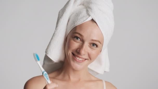 Attraente ragazza allegra con asciugamano sulla testa e spazzolino da denti in mano felicemente guardando in macchina fotografica isolata - Filmati, video