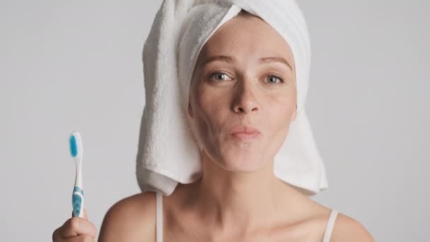Mladá legrační žena s ručníkem na hlavě a zubní kartáček v ruce s úsměvem ukazující čisté zuby na kameře izolované - Záběry, video