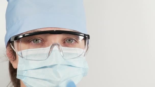 femme dans un masque médical jetable, lunettes et une casquette textile regarde la caméra sur un fond blanc, les yeux fatigués - Séquence, vidéo