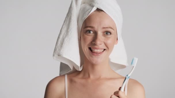 Aantrekkelijk positief meisje met handdoek op hoofd en tandenborstel in de hand vreugdevol kijkend in camera het controleren van schoon van tanden over grijze achtergrond - Video