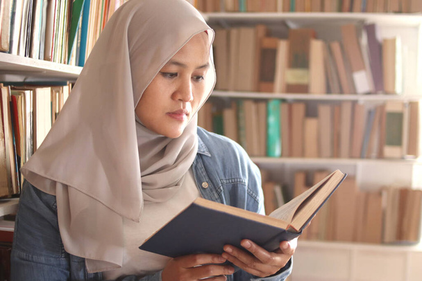 Asian muslim woman wearing hijab reading book in library, educational concept. Happy smiling expression when doing leisure activity - Фото, изображение