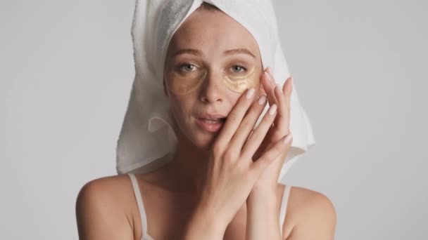 Mladá krásná žena s ručníkem na hlavě nanášení zlaté oční pásky šťastně dívá ve fotoaparátu izolované. Krása konceptu - Záběry, video