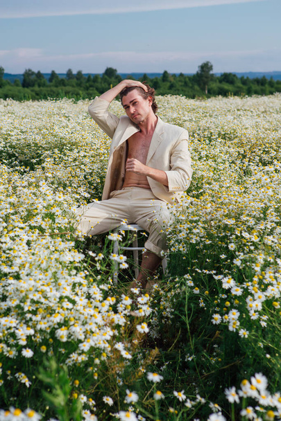 Tall handsome man dressed in a white suit on on naked body sitting on a white chair in camomile flowers field - Photo, Image