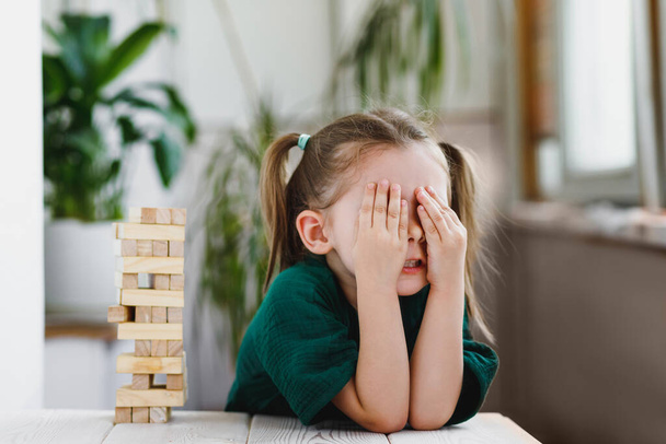 Cute caucasian little girl with ponytails closing her face with her hands, a wooden jenga tower standing on a table. Selective soft focus - Foto, afbeelding