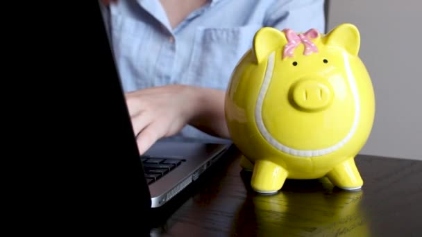Close-up niedlichen gelben Sparschwein voller Münzen. Frau tippt auf ihrem Computer mit einem Sparschwein mit Geld im Vordergrund. Bürostil. Raum für Text. - Filmmaterial, Video