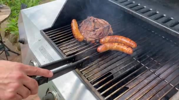 Lifestyle summer outdoor man cooking stakes and sausages on a propane gas grill holing metal tongues - Footage, Video