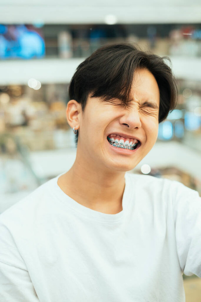 Tiener man in wit t-shirt heeft een grote heldere glimlach op zijn gezicht terwijl hij wacht op zijn vriend in het winkelcentrum. - Foto, afbeelding