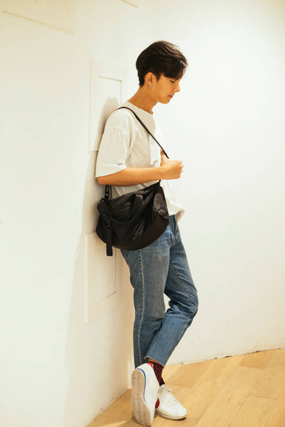 Teenage guy leaning againt the white wall looking at the wooden floor. - 写真・画像