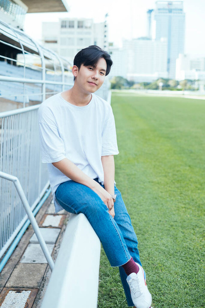 Adolescente em jeans e t-shirt branca sentado na barricada no campo de futebol
. - Foto, Imagem