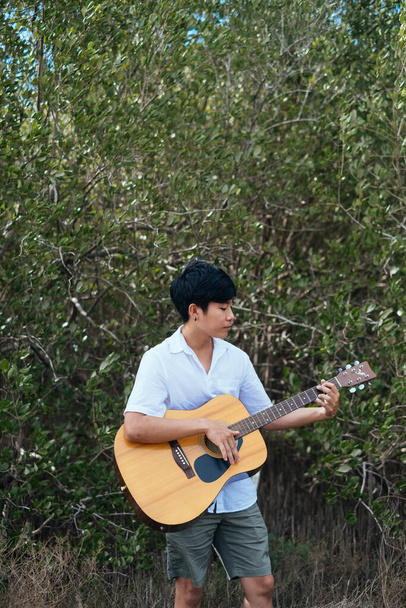 Музыкант в белой рубашке стоит перед мангровым лесом, держа гитару на руках. - Фото, изображение