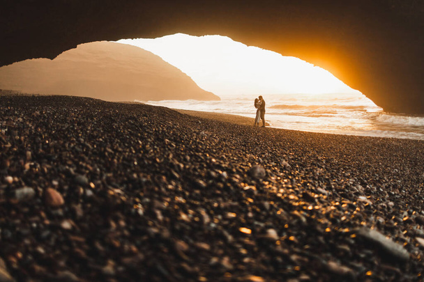 Schönes Paar, das sich unter einem großen Bogen am Strand von Legzira küsst und einen fantastischen warmen Sonnenuntergang genießt. Berühmtes und beliebtes Wahrzeichen Marokkos. Schönes Abendlicht. - Foto, Bild