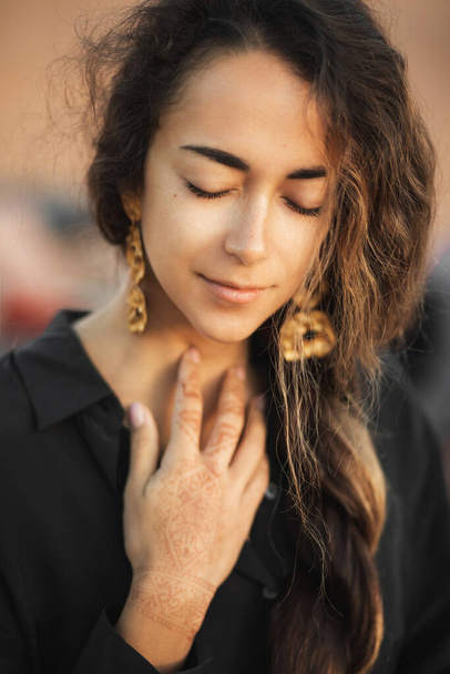 Close up portrait of young oriental brunette woman with long curly hair and closed eyes. Black shirt and golden earrings. Pretty female face and henna tattoo on hand. - 写真・画像