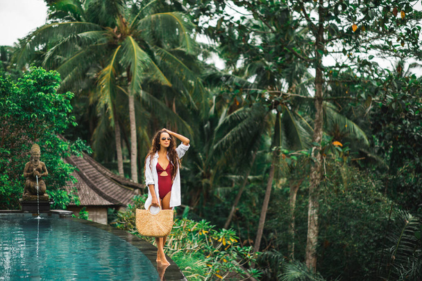 Woman in sunglasses, red swimsuit and white shirt relaxing and walking by poolside. Handmade straw bag. Infinity swimming pool in jungle. - Photo, Image