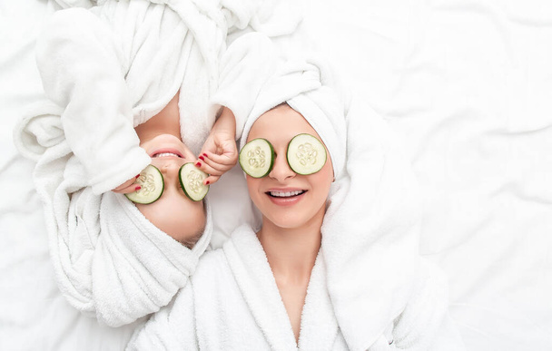 Positive mother and daughter doing face mask applying pieces of cucumber to their eyes, wrapped in a white bath towel. Long banner format, top view - Photo, Image