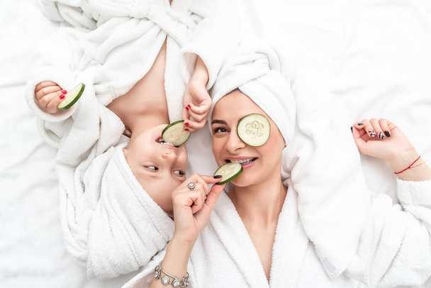 Madre e hija positivas haciendo mascarilla facial aplicando trozos de pepino a sus ojos, envueltas en una toalla de baño blanca. vista superior
, - Foto, Imagen