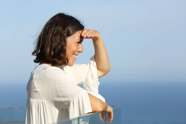Side view portrait of a happy adult woman looking at views protects from sun standing in a balcony on the beach - Фото, зображення