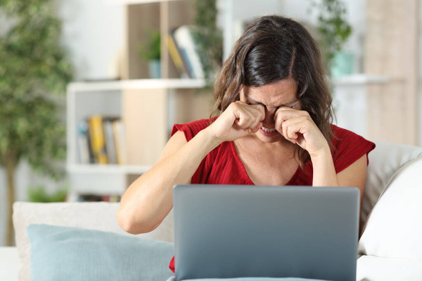 Adult woman with laptop scratching itchy eyes sitting in the sofa in the living room at home - Foto, imagen