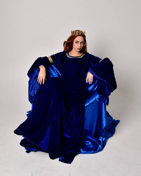 Full length portrait of  girl wearing long blue velvet gown with golden crown. sitting pose, isolated against a studio background. - Photo, Image