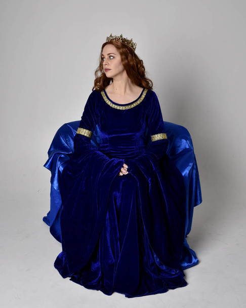 Retrato de comprimento total da menina vestindo longo vestido de veludo azul com coroa de ouro. postura sentada, isolada contra um fundo de estúdio
. - Foto, Imagem