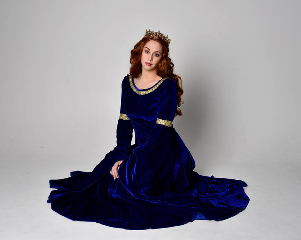 Full length portrait of  girl wearing long blue velvet gown with golden crown. sitting pose, isolated against a studio background. - Foto, immagini