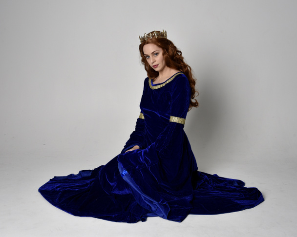 Portrait pleine longueur de fille portant une longue robe de velours bleu avec couronne dorée. pose assise, isolée sur fond de studio. - Photo, image