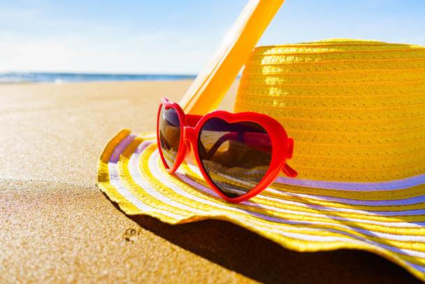 Sun protection accessories on sea shore. Red sunglasses, yellow straw hat and sunscreen cream bottle on sand beach. Summer and holidays. Skincare. - Foto, Imagen