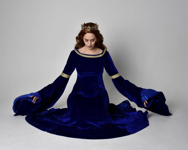 Портрет девушки в длинном синем бархатном платье с золотой короной. сидячая поза, изолированная на фоне студии. - Фото, изображение
