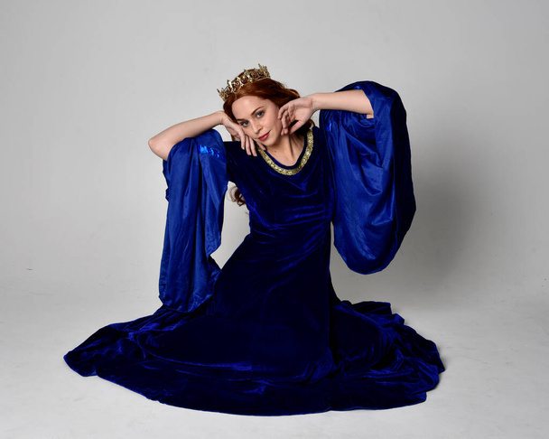 Portrait pleine longueur de fille portant une longue robe de velours bleu avec couronne dorée. pose assise, isolée sur fond de studio. - Photo, image