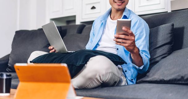 Mladý usměvavý asijský muž relaxaci pomocí notebooku pracovní a video konference setkání doma. Mladý kreativní muž při pohledu na obrazovce psaní zprávy s smartphone.work z domova koncept - Fotografie, Obrázek