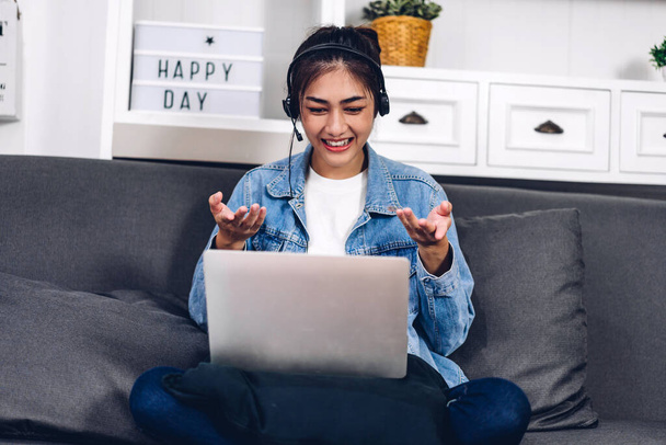 Genç gülümseyen mutlu Asyalı kadın dizüstü bilgisayarı kullanarak rahatlıyor ve evde video konferansı yapıyor. Genç yaratıcı kız kulaklıkla konuşuyor. Ev konseptinden çalışma. - Fotoğraf, Görsel