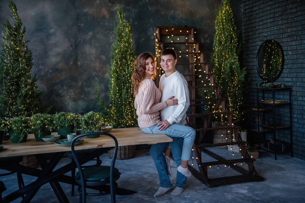 A beautiful girl with curls sits on a wooden table, she is hugged by a young man, dressed in winter warm sweaters and jeans, against the background of trees and lights. loving couple hugging and looking at the camera. Harmonious relationship concept - Φωτογραφία, εικόνα