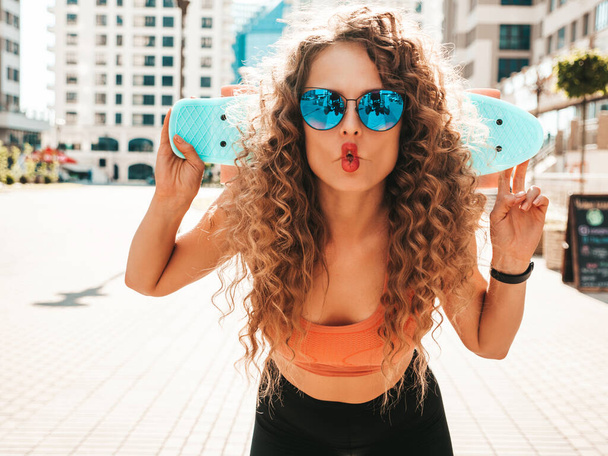 Young beautiful sexy smiling hipster woman with afro curls hairstyle.Trendy girl in summer clothes.Positive female with blue penny skateboard posing on the street background.Makes fish face - Foto, Imagen