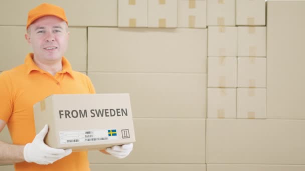 Warehouse worker holds cardboard box with printed FROM SWEDEN text on it - Záběry, video