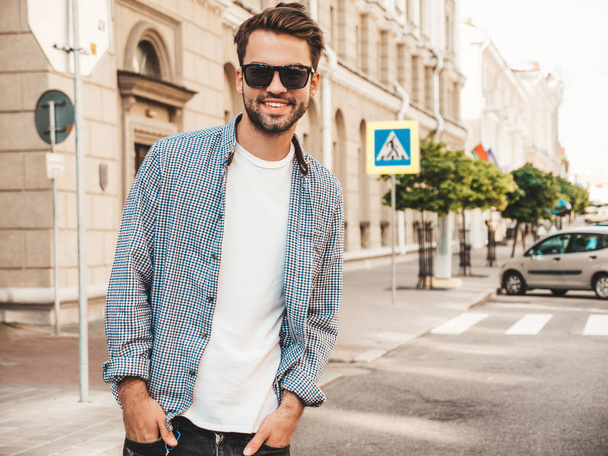 Portrait of handsome smiling stylish hipster lambersexual model.Man dressed in white T-shirt. Fashion male posing on the street background in sunglasses - 写真・画像