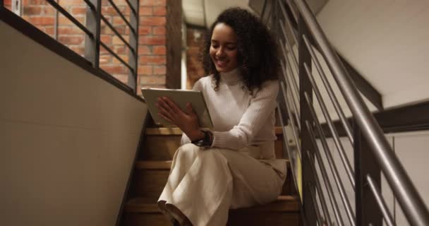 Mixed race professional businesswoman working in a modern office using digital tablet, sitting on staircase, smiling. Business creativity technology. - Filmagem, Vídeo